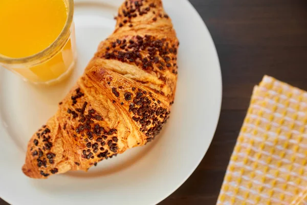 Croissant fresco em uma placa cerâmica e toalha de cozinha com uma xícara de suco de laranja. Croissant recém-assado em uma mesa de madeira escura . — Fotografia de Stock