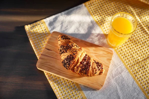 Croissant fresco en una tabla de madera y una toalla de cocina con una taza de jugo de naranja. Croissant recién horneado sobre una mesa de madera oscura . Imagen de stock