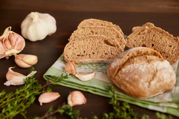 Skład z czosnkiem i chleb na ciemny drewniany stół. Świeżo upieczony chleb ręcznie robione na Ręcznik kuchenny. Chleb jest pokroić w plasterki i cały bochenek jest nie daleko. — Zdjęcie stockowe
