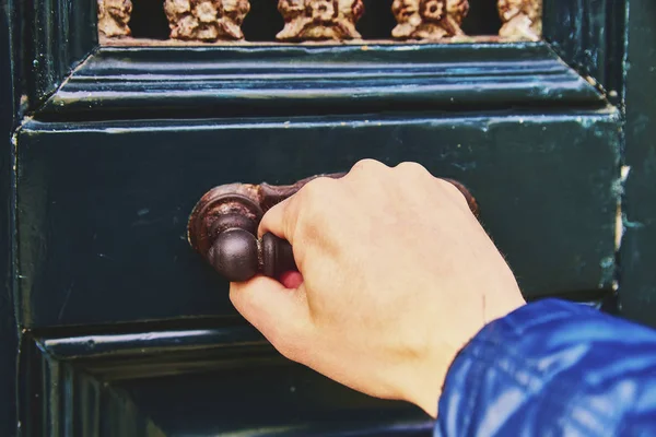 A man\'s hand opens the door, a close-up photo. The hand holds onto the antique metal handle of the green-blue door.