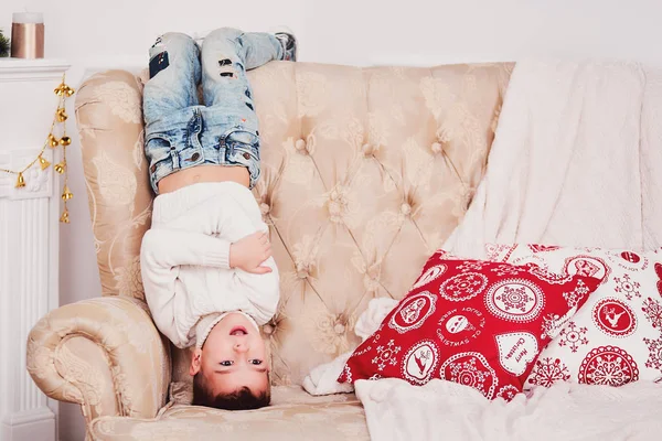 A cute boy is standing on his head on the couch. A sporty guy in a white knitted sweater and jeans and a stylish hairdo stands upside down on the sofa. Near the pillow with a red Christmas pattern.