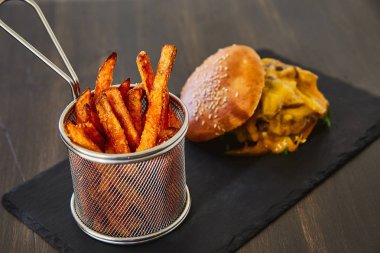 Susam, eritilmiş sarı peynir, yeşil taze salata ve koyu ahşap bir masa üzerinde bir kara tahta üzerinde taze chop ile toplanan bir topuz çizburger. Parlak metal kavanozda büyük tuz ile kızarmış patates ile.