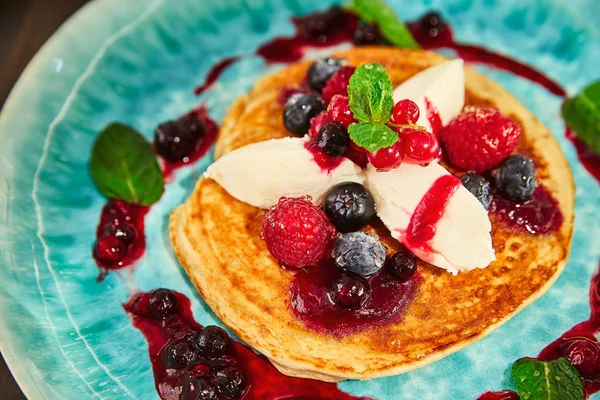 A large homemade pancake with white mascarpone cream, raspberries, blueberries, currants, berries and honey on a blue plate. It is decorated with jam from mountain ash and green young leaves of mint.