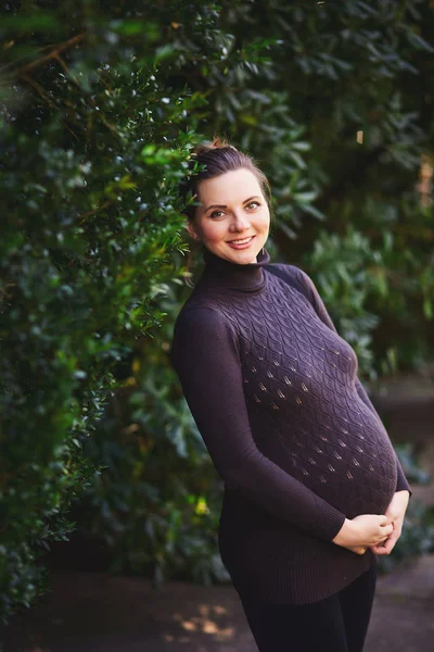 Портрет улыбающейся красивой беременной женщины с сверкающим взглядом в вязаном свитере, держащемся за живот в парке. Симпатичная девушка стоит в саду на фоне зеленой листвы . — стоковое фото