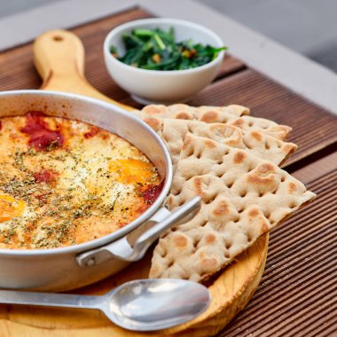 Domates, peynir ve ev yapımı ince lavaş ve ahşap bir kahverengi masa üzerinde sulu yeşillik ile kulplu bir alüminyum tava içinde baharatlar ile omlet. Doğal ışık altında fotoğraf çekildi.