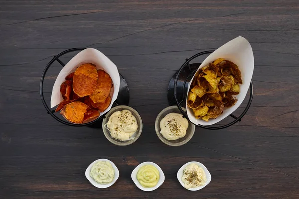 Um conjunto de muitos lanches para vinho ou cerveja: dois tipos de batatas fritas caseiras e cinco molhos diferentes em uma mesa de madeira marrom. Vista de cima, vista superior . — Fotografia de Stock