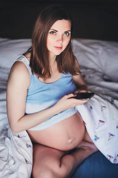 Die junge attraktive Schwangere sitzt mit dem Handy in der Hand auf dem Bett. schöne schwangere Frau in einem hochgekrempelten Tanktop mit nacktem Bauch auf dem Bett sitzend und ein Handy in der Hand. — Stockfoto