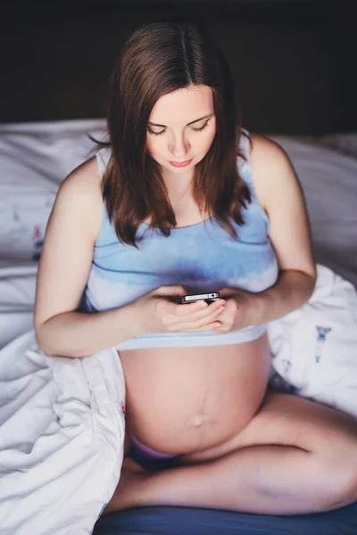 Genç çekici hamile kadın elinde cep telefonu ile yatakta oturuyor. Toplu tank top yatağın üzerine oturan ve bir cep telefonu tutan çıplak bir göbek ile bir güzel hamile kadın. — Stok fotoğraf