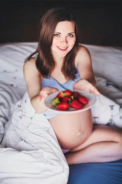 Attraktive junge schwangere Frau sitzt auf einem Bett und reicht einen Teller Erdbeeren. Nahrung für eine glückliche Schwangere, Obst und Gemüse für eine ausgewogene Ernährung der zukünftigen Mutter. — Stockfoto
