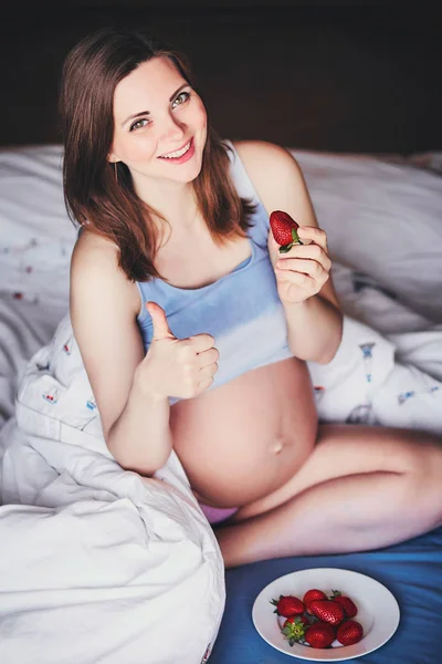 Attraktive junge schwangere Frau sitzt auf einem Bett und isst eine reife Erdbeere. Nahrung für ein glückliches schwangeres Mädchen, Obst und Gemüse für eine ausgewogene Ernährung zukünftige Mutter. Gesunde Ernährung für eine erfolgreiche Schwangerschaft. — Stockfoto