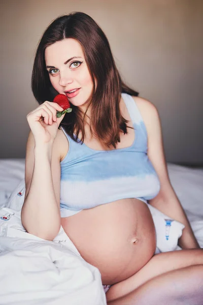 Attraktive junge schwangere Frau sitzt auf einem Bett und isst eine reife Erdbeere. Nahrung für ein glückliches schwangeres Mädchen, Obst und Gemüse für eine ausgewogene Ernährung zukünftige Mutter. Gesunde Ernährung für eine erfolgreiche Schwangerschaft. — Stockfoto
