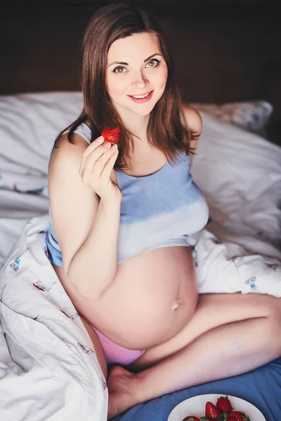Nahrung für ein glückliches schwangeres Mädchen, Obst und Gemüse für eine ausgewogene Ernährung zukünftige Mutter. attraktive junge schwangere Frau sitzt auf einem Bett und isst eine reife Erdbeere. Gesunde Ernährung für eine erfolgreiche Schwangerschaft. — Stockfoto