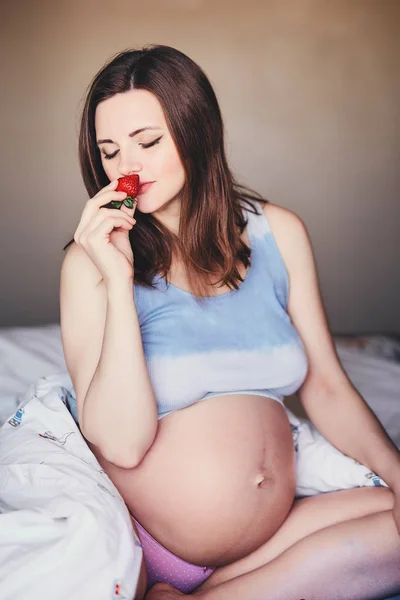 Привлекательная молодая беременная женщина сидит на кровати и нюхает спелую клубнику. Еда для счастливой беременной девочки, фрукты и овощи для сбалансированного питания будущей мамы. Здоровое питание для успешной беременности . — стоковое фото