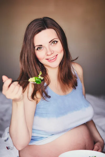 Беременная улыбающаяся девушка в футболке сидит на кровати и ест салат с зелеными и красными листьями. Молодая женщина соблюдает диету во время беременности. Здоровое питание для будущих матерей. Счастливая женщина наслаждается беременностью . — стоковое фото