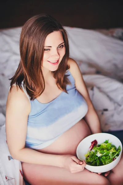 Schwangere lächelnde Mädchen im T-Shirt sitzen auf dem Bett und essen Salat mit grünen und roten Blättern, trinken Orangen-, Kürbis- und Karottensaft. junge Frau halten Diät während der Schwangerschaft. Gesunde Ernährung für werdende Mütter. — Stockfoto