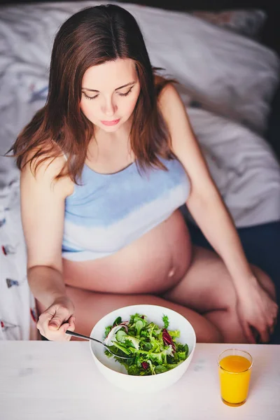 A menina grávida em uma camiseta senta-se na cama e come salada com folhas verdes e vermelhas, bebe suco de laranja, abóbora e cenoura. Mulher jovem manter a dieta durante a gravidez. Alimentação saudável para gestantes . Fotos De Bancos De Imagens