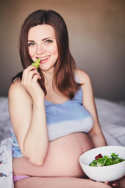 T-shirt hamile gülümseyen kız yatağa otur ve yeşil ve kırmızı yaprakları salata yiyin. Genç kadın gebelik sırasında diyet tutmak. Sağlıklı gıda bekleyen anneler için. Mutlu kadın hamile zaman zevk.