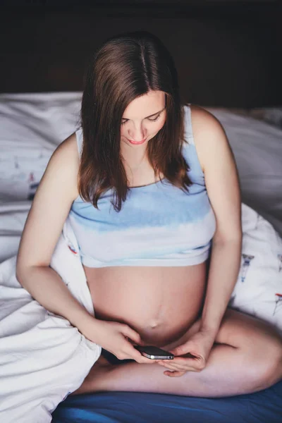 Genç çekici hamile kadın elinde cep telefonu ile yatakta oturuyor. Toplu tank top yatağın üzerine oturan ve bir cep telefonu tutan çıplak bir göbek ile bir güzel hamile kadın. — Stok fotoğraf