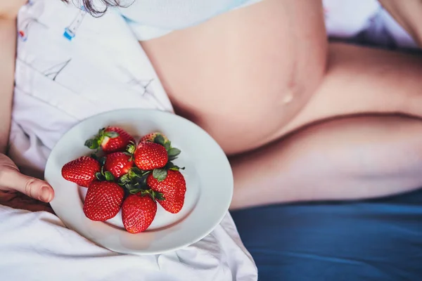 Çekici genç hamile kadın bir yatakta oturur ve olgun çilek yiyor. Gıda mutlu hamile kız, meyve ve sebzeler için dengeli beslenme gelecekteki anne. Başarılı bir gebelik için sağlıklı gıda. — Stok fotoğraf