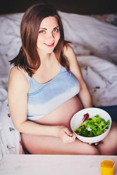 Schwangere lächelnde Mädchen im T-Shirt sitzen auf dem Bett und essen Salat mit grünen und roten Blättern, trinken Orangen-, Kürbis- und Karottensaft. junge Frau halten Diät während der Schwangerschaft. Gesunde Ernährung für werdende Mütter. — Stockfoto