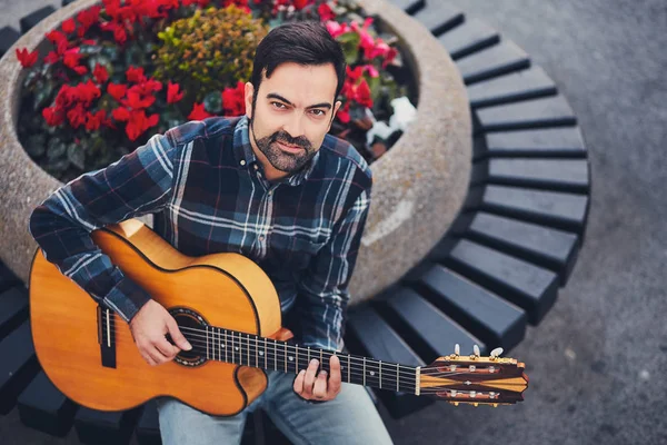 Homem elegante com barba em camisa xadrez e jeans na rua perto de um parque com flores e uma guitarra em suas mãos. Macho com um belo sorriso senta-se à noite no banco e toca guitarra . — Fotografia de Stock