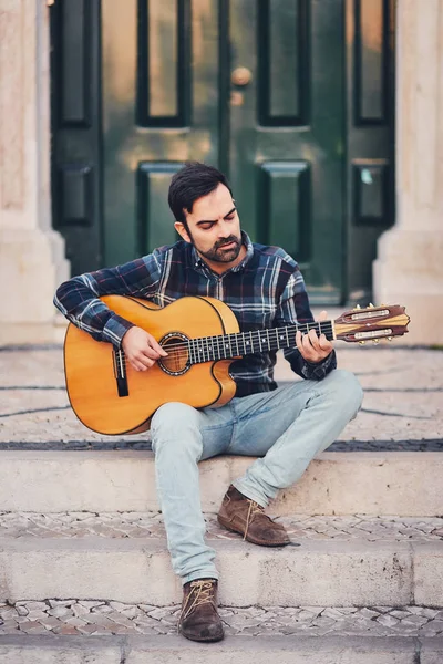 Homem elegante com barba em camisa xadrez e jeans na rua nos degraus perto da porta. Macho com um belo sorriso senta-se à noite no banco e toca guitarra. Estilo moderno de vida . — Fotografia de Stock