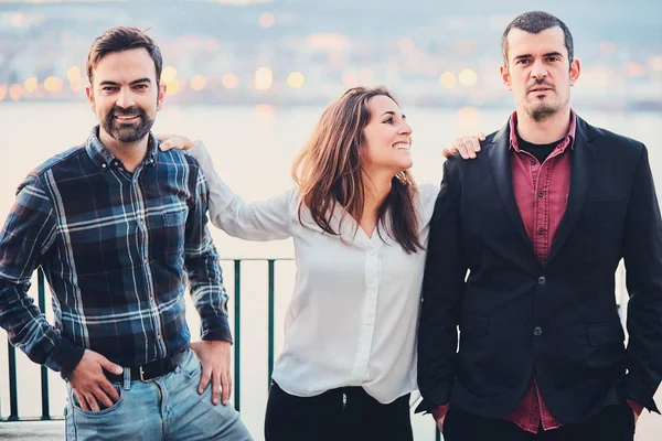 Two men and one woman are standing next to smile and joke, have fun. Friends laugh at the background of the evening city and the river. Modern youth lifestyle.