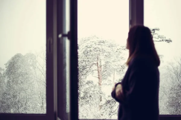The girl is standing at the window and looks at how it\'s snowing