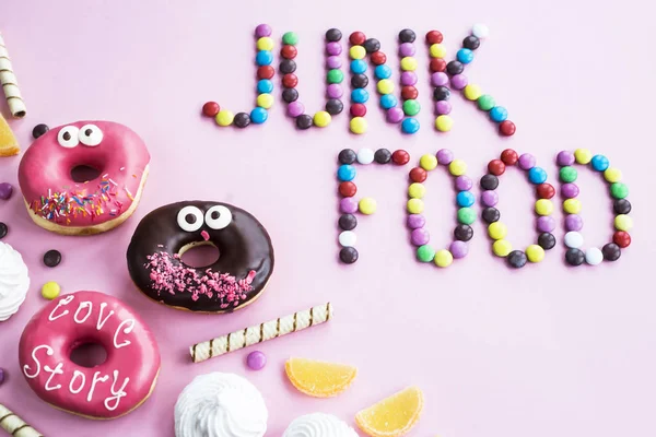 The phrase Junk food is lined with chocolate glazed balls on a pink background. Next to donuts, marmalade, chocolate sticks and meringue. Unhealthy food concept. Top view