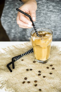 A girl in a gray dress inserts a straw into a glass of cooked iced coffee with milk. Summer cooling drink concept clipart