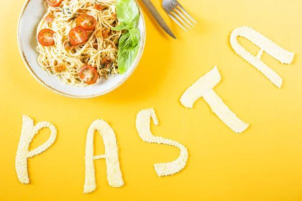 The word pasta is laid out of small pasta next to a plate with head spaghetti with cherry and basil. Top view, flat lay