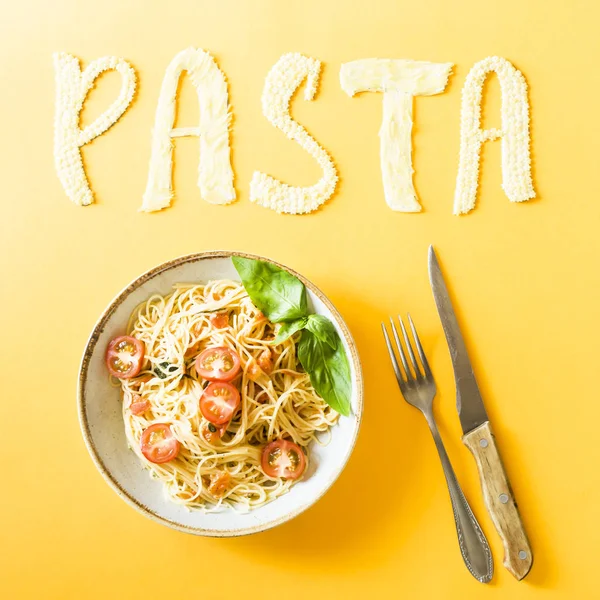 The word pasta is laid out of small pasta next to a plate with head spaghetti with cherry and basil. Top view, flat lay