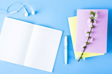 Açık defter ile kulaklık, bir fincan çay ve mavi bir arka plan üzerinde renkli not defterleri. Çiçekli dallar ile bahar işyeri