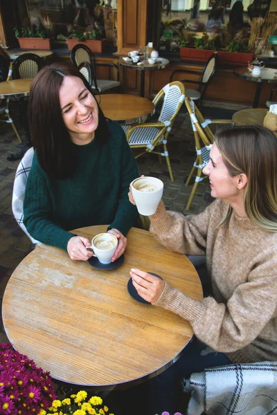 Two joyful female friends are drinking cappuccino in a city street coffee shop.