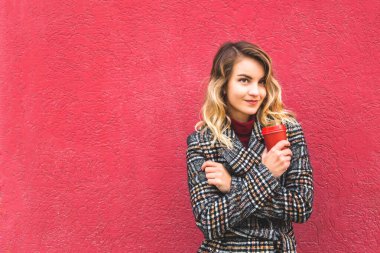 Güzel beyaz kız elinde sarı bir sonbahar yaprağı ve kırmızı duvarın yanında duran kırmızı bir fincan kahve tutuyor..