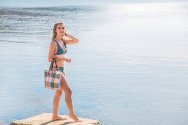 Millennial sarışın kadın elinde bir kumsal çantasıyla deniz kenarında geziyor ve dinleniyor..