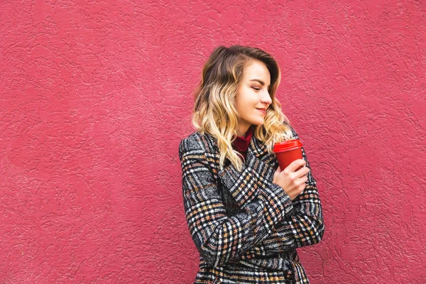 Όμορφο καυκάσιο κορίτσι που κρατά ένα κίτρινο φύλλο του φθινοπώρου και ένα κόκκινο φλιτζάνι καφέ στέκεται κοντά στον κόκκινο τοίχο. — Φωτογραφία Αρχείου