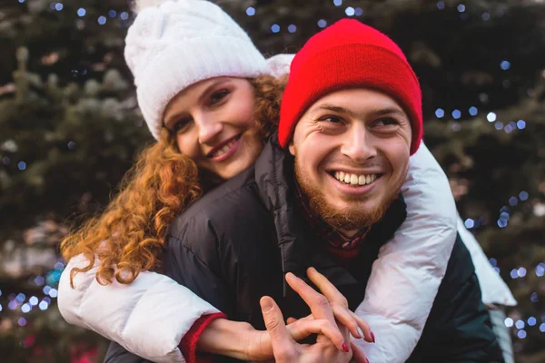 Der Kerl und das Mädchen in den roten Pullovern lächeln glücklich am Weihnachtsbaum. — Stockfoto