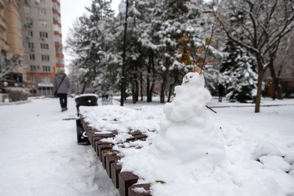 little funny snowman on a bench in a city park on a background of snowy trees. snow woman. New Year\'s holidays. winter fun