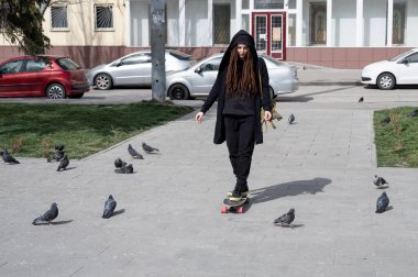 Siyah mantolu rastalı bir kız güvercinlerin arasında uzun bir tahtaya biniyor. Üzgün kız şehirde dolaşıyor.