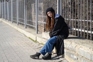 Siyah şapkalı, siyah ceketli, rastlantısal bir kız. Modern moda grunge 'da. Kaldırımda oturan kız.