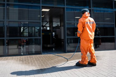 Turuncu üniformalı bir adam binaya giriş yolunu dezenfekte ediyor. Viral enfeksiyonun yayılmasını önlemek için. açık alan temizliği