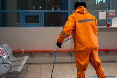 Turuncu giysili bir adam tren istasyonundaki salona kimyasal püskürtüyor. Halka açık yerlerin dezenfekte edilmesi. Koronavirüsün yayılmasını engelliyor.