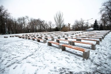 Sahnenin önündeki şehir parkında karla kaplı banklar. Karda insanların ayak izleri. Şehirde soğuk hava ve boş bir park.