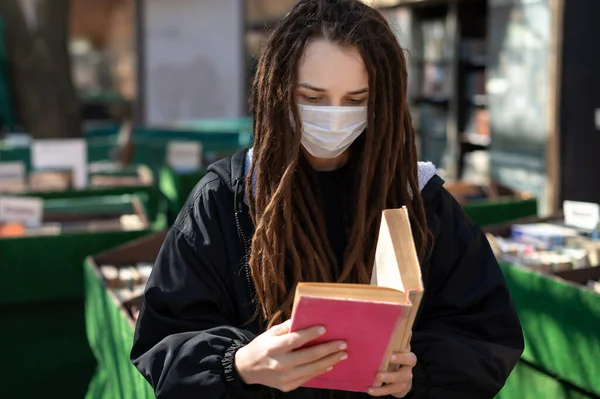 girl with dreadlocks in a medical mask is reading a book. coronavirus infection. Young woman wearing protective mask on face