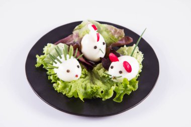 Hayvana benzeyen yumurtalar (biberiye, turp, soğan gibi elementlerle) bir tabakta, beyaz arkaplanda.