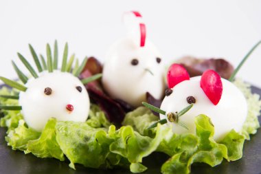 Hayvana benzeyen yumurtalar (biberiye, turp, soğan gibi elementlerle) bir tabakta, beyaz arkaplanda.