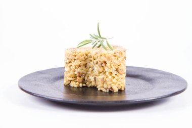 Kalp şeklinde Risotto porsiyonu, düğme mantarlı ve pastırmalı. Beyaz arka planda siyah bir tabakta biberiye dalıyla süslenmiş.