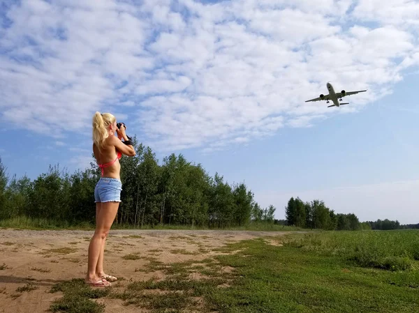 Blond flicka med långt hår tar bilder av ett plan som flyger över henne på en molnig himmel bakgrund — Stockfoto