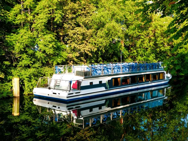 Pleasure boat is reflected in the water of the canal, surrounded by green foliage Stockfoto